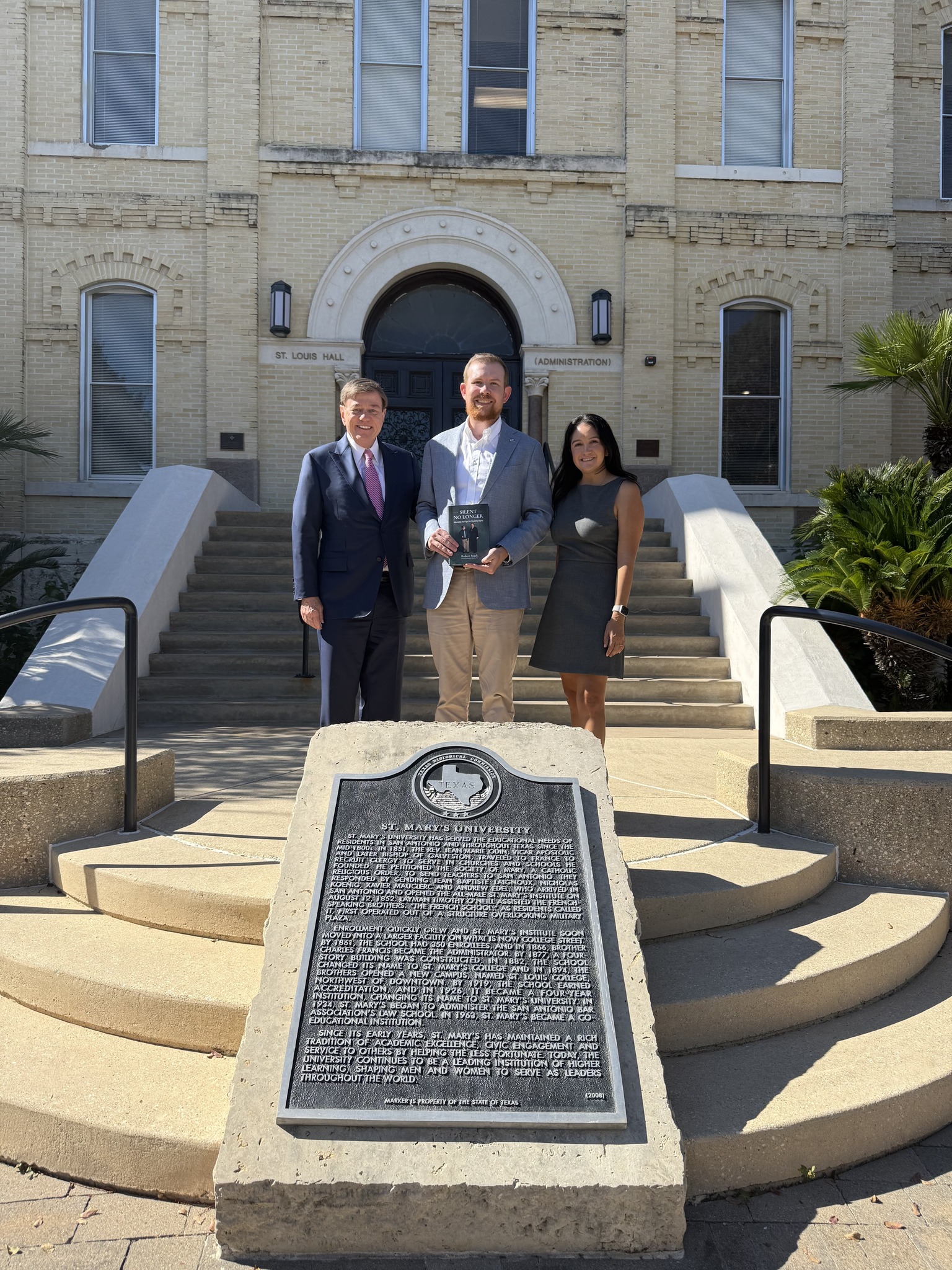 Robert Stack stands outdoors with Dr. Doty and Samantha Cutler in front of St. Louis Hall at St. Mary’s University; Dr. Doty holds a copy of Silent No Longer as the group poses in sunlight near a Texas Historical Commission plaque.