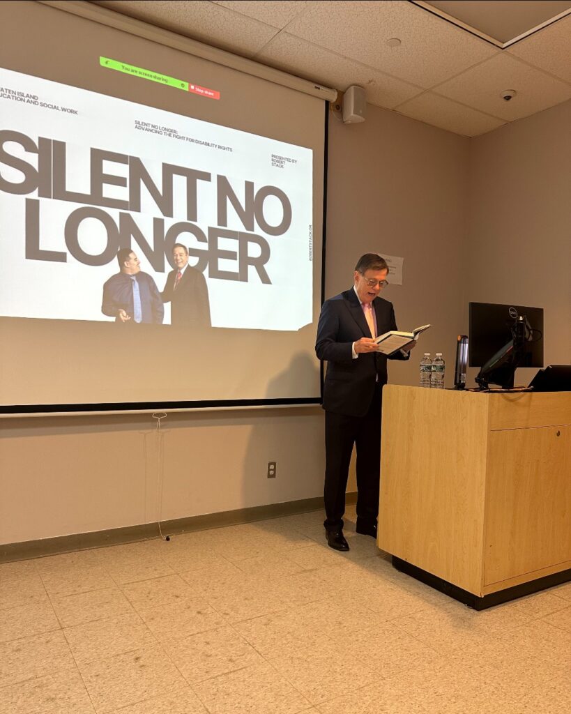 Robert Stack stands behind a podium reading from “Silent No Longer” during a guest lecture, with a presentation slide from the book projected behind him at the College of Staten Island.