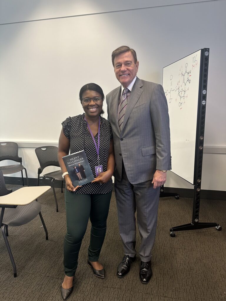 Robert Stack stands beside Professor Dora Onwumere at New York University; she holds a copy of “Silent No Longer” as they pose together indoors following a guest lecture.