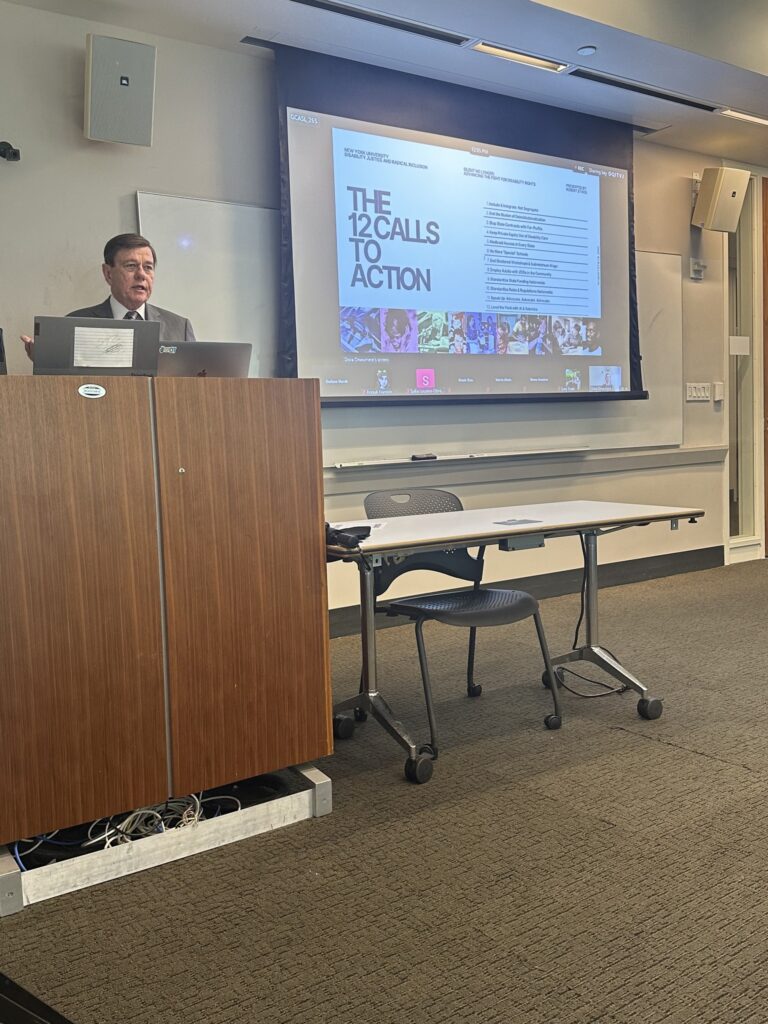 Robert Stack speaking behind a podium at New York University with a presentation slide titled “12 Calls to Action” displayed during his lecture.