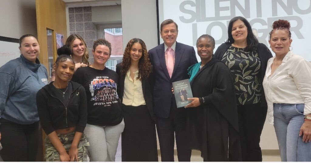 Nine people pose together indoors after a guest lecture at the College of Staten Island; one person holds a copy of “Silent No Longer.” A presentation slide from the talk is visible in the background.